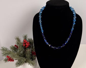 Può includere: Una collana di perline con un gradiente di perle di vetro blu in varie tonalità, che vanno dall'azzurro cielo al blu navy intenso. La collana è composta da perle rotonde e sfaccettate alternate a piccoli distanziatori color argento.