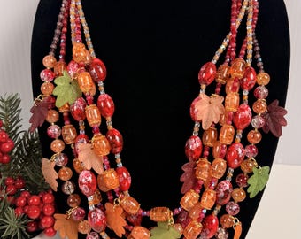 Può includere: Una collana di perline a più fili caratterizzata da una varietà di perline rosse, arancioni e color ambra intervallate da piccoli ciondoli a forma di foglia nei toni del verde, marrone e marrone chiaro, dall'aspetto stratificato.