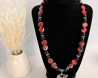 Può includere: Una collana con perline rosse e nere alternate e distanziatori color argento. Le perline rosse presentano un motivo marmorizzato, mentre quelle nere sono lisce e rotonde. Un ciondolo circolare color argento con un intricato design traforato pende al centro.