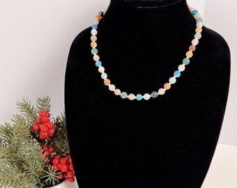 Può includere: Una collana di perline composta da piccole pietre rotonde multicolori nei toni del blu, arancione, verde, rosa e bianco, separate da minuscoli distanziatori dorati. La collana è esposta su un busto nero su sfondo neutro.