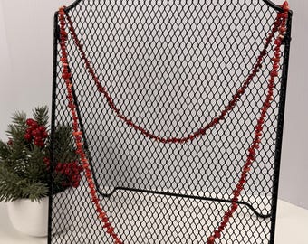 Può includere: Una collana lunga composta da piccole pietre rosse irregolari a forma di scaglie. La collana è appesa su un espositore in filo metallico nero con una rete a rombi e una parte superiore arcuata decorativa con un anello circolare.
