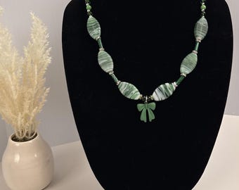 Può includere: Una collana con perline ovali marmorizzate verdi e bianche, alternate a piccoli distanziatori color argento e perline rotonde verde scuro. Al centro della collana è presente un ciondolo verde opaco a forma di fiocco appeso a un anello argentato.