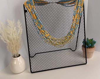 Può includere: Una collana a più fili con piccole perline rotonde in tonalità alternate di giallo brillante e azzurro. La collana è esposta su un supporto in rete metallica nera, che mette in mostra diversi fili sovrapposti che cadono a forma di U.