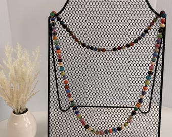 Può includere: Una collana lunga caratterizzata da un filo continuo di piccole perline rotonde multicolori nelle tonalità del rosso, blu, verde, giallo e nero, drappeggiata su un espositore in filo metallico nero con parte superiore curva e griglia.