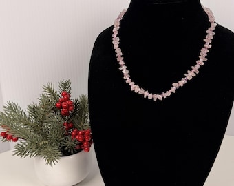 Può includere: Una collana composta da piccole pietre rosa irregolari a forma di scaglie infilate insieme. La collana è esposta su un busto nero, mettendo in risalto l'aspetto strutturato e grezzo dei singoli pezzi di pietra rosa disposti in un cerchio continuo.