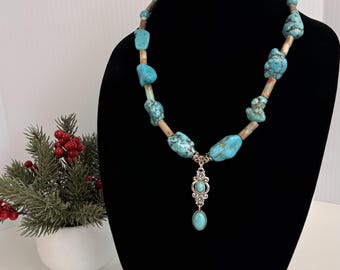 Può includere: Una collana con pietre di turchese irregolari e levigate, alternate a perline cilindriche color sabbia. Il pezzo presenta un ciondolo centrale in metallo argentato con dettagli in filigrana e una goccia di turchese ovale.