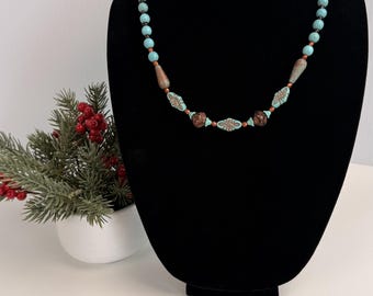 Può includere: Una collana di perline composta da perline rotonde color turchese, perline marroni allungate e affusolate, e perline turchesi piatte di forma ovale con motivi marroni incisi, intervallate da piccole perline distanziatrici color rame.