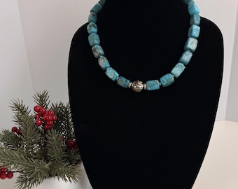 Può includere: Una collana composta da perline rettangolari in pietra color turchese con venature naturali. Le perline sono infilate insieme e presentano una singola perlina rotonda in metallo argentato testurizzato posizionata al centro.