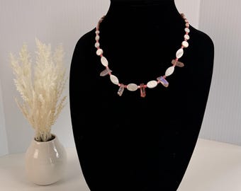 Può includere: Una collana di perline composta da un'alternanza di perline bianche di forma ovale e piccoli frammenti traslucidi simili a cristalli dai toni rosati. Le perline sono infilate su un sottile cordino rosa, creando un motivo circolare sul supporto.