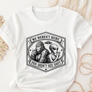 Op de afbeelding: Een wit T-shirt met korte mouwen en een zwarte grafische print van Bigfoot en een alien die samen roken. Boven hen staat een vliegende schotel en de tekst WE WERENT HERE, en daaronder de tekst YOU DIDNT SEE SHIT, alles binnen een kader.