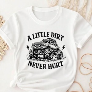 May include: A white short-sleeved t-shirt featuring a black graphic print of a monster truck with large tires. The text A LITTLE DIRT NEVER HURT is printed in a bold, arched font above and below the truck, accompanied by two lightning bolt icons.