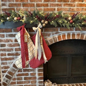 以下が含まれることがあります： 暖炉の棚に吊るされた2つのクリスマスストッキング。1つは黒い格子模様のクリーム色で赤いリボンが付いており「Daddy」と書かれています。もう1つは無地の赤でストライプのリボンが付いており「Mommy」と書かれています。上部には松の枝とベリーのガーランドが飾られています。