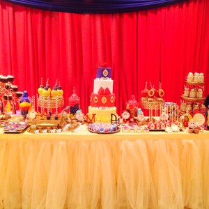 May include: A dessert table with a yellow tulle skirt, red backdrop, and blue drape. The table is laden with cupcakes, cake pops, a multi-tiered cake, and other treats. A Snow White doll is on the right.
