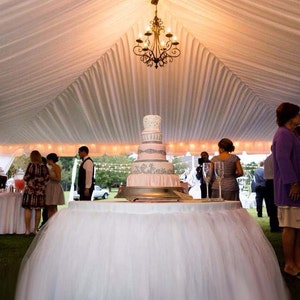 May include: A white wedding cake with pink and gray frosting and silver embellishments sits on a table with a white tulle tablecloth. A chandelier hangs from the white tent ceiling above the cake.