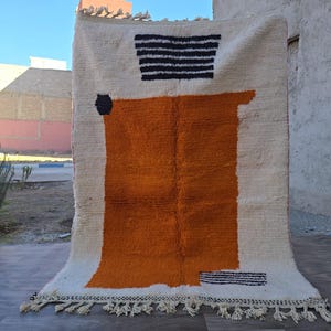 May include: A rectangular area rug featuring a cream-colored base with a large, solid orange block in the center. The design includes a black and white striped trapezoid at the top, a small black hexagon on the left, and black fringe along the bottom edge.
