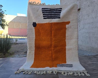 May include: A rectangular area rug featuring a cream-coloured base with a large, solid orange block in the centre. The design includes a black and white striped trapezoid at the top, a small black hexagon on the left, and black fringe along the bottom edge.