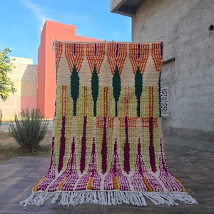 May include: A rectangular wool rug featuring a cream-colored base with a geometric pattern in orange, dark green, magenta, and yellow. The design consists of vertical diamond and chevron shapes, finished with a white knotted fringe at the bottom edge.