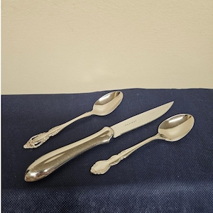 May include: A set of three silver-toned utensils consisting of two spoons and one knife. The spoons feature ornate, embossed detailing on the handles, while the knife has a smooth, plain handle and a blade with the engraved text Oneida.