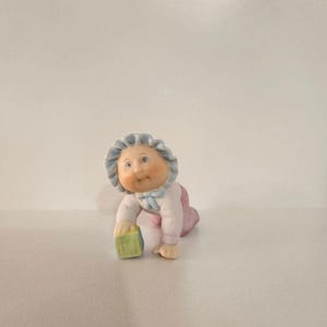May include: A small ceramic figurine of a crawling baby wearing a light pink outfit and a ruffled blue bonnet. The baby has rosy cheeks and is holding a pale green square block with a letter embossed on the front, captured in a crawling pose.