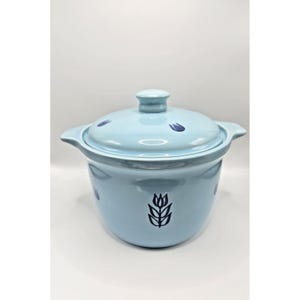 May include: A light blue ceramic casserole dish with a matching lid, featuring dark blue tulip motifs on the body and lid. The pot has two side handles and a central knob on the lid, with a smooth, glossy finish throughout the exterior.