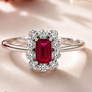 May include: A silver ring featuring a central emerald-cut red gemstone surrounded by a halo of ten small round clear stones. The text YAKUTTA JEWELLERY & ACCESSORIES 925 SILVER is printed below the ring with a diamond-shaped logo icon.