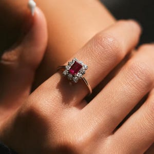 Puede incluir: Un anillo con una piedra preciosa roja rectangular de corte esmeralda en el centro, rodeada por un halo de peque&ntilde;as piedras blancas redondas. El engaste es de metal plateado pulido y la banda es delgada y lisa, rodeando el dedo.