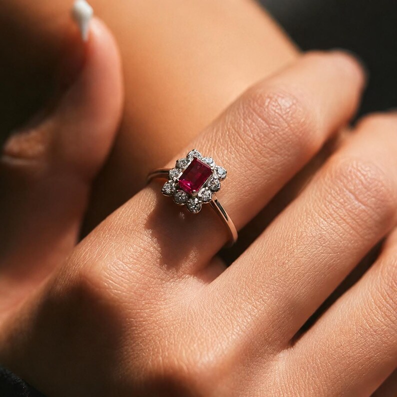 Puede incluir: Un anillo con una piedra preciosa roja rectangular de corte esmeralda en el centro, rodeada por un halo de peque&ntilde;as piedras blancas redondas. El engaste es de metal plateado pulido y la banda es delgada y lisa, rodeando el dedo.