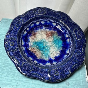 May include: A handcrafted, round, blue ceramic plate with a textured, speckled finish. The plate features a scalloped edge and a central design of fused glass in shades of blue, white, and brown. Decorative floral-like patterns adorn the rim.