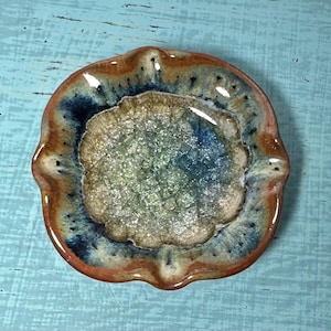 May include: A handmade ceramic ashtray with a unique, organic shape. The ashtray features a central area with a textured, crackled appearance, surrounded by a rim with a blend of brown, blue, and cream colors. The overall design is artistic and decorative.