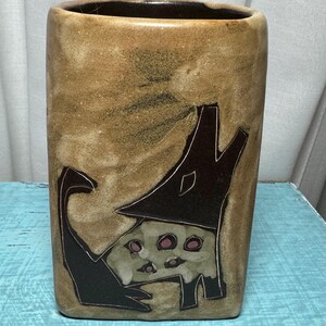 May include: A rectangular ceramic vase with a tan background and a dark brown rim. The vase features a stylized illustration of a dog wearing a hat. The dog is depicted in dark brown and tan with pink accents. The vase is likely handmade.