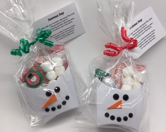 May include: Two cellophane bags filled with marshmallows, candies, and a snowman-themed treat. Each bag is decorated with a white paper snowman face and a tag that reads "Snowman Soup". The bags are tied with green and red ribbons.