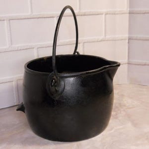 May include: A black cast iron cauldron with a curved handle. The cauldron has a spout and two small decorative protrusions on the side. The background is a white brick wall.