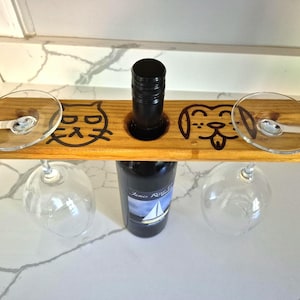May include: A rectangular wooden wine glass holder with a central circular cutout for a bottle neck. The wood features a dark brown engraved cat face on the left and a dog face on the right, with two slots to hold the stems of two clear glass wine glasses.