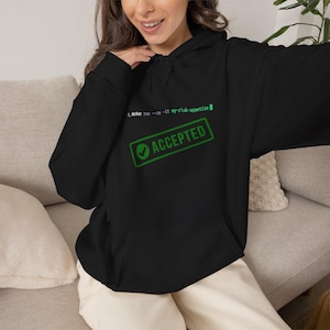 Op de afbeelding: Een zwarte hoodie met een groene grafische print op de borst. Het ontwerp toont de tekst docker run --rm -it my-risk-appetite boven een rechthoekige stempelafbeelding met een vinkje en het woord ACCEPTED in het groen.