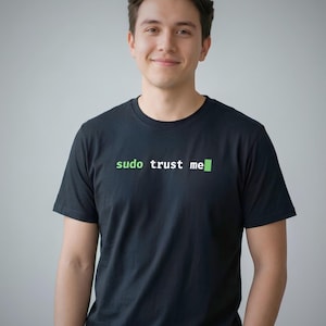 以下が含まれることがあります： 胸元に白と緑で「sudo trust me」というテキストがプリントされた黒の半袖Tシャツ。単語「sudo」と末尾のブロックカーソルは緑色で、「trust me」という単語は白色で、すべて等幅フォントでデザインされています。
