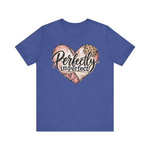 以下が含まれることがあります： ヘザーブルーの半袖Tシャツ。中央にはピンクとベージュの色合いで縫い合わされたパッチワーク風のハートのグラフィックが描かれています。ハートには黒い筆記体で「Perfectly imperfect」という文字と、パッチ部分に小さなハートのアイコンがあしらわれています。