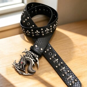 Puede incluir: Un cinturón negro con un patrón texturizado de piel de serpiente, con una hebilla de metal plateado en forma de dragón con un ojo rojo. La correa está decorada con tachuelas de metal plateado y emblemas de flor de lis a lo largo de la pieza.
