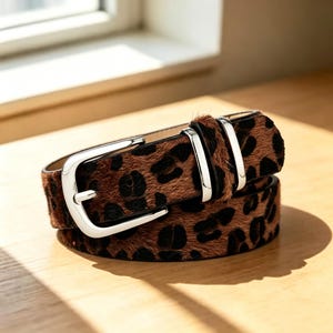 May include: A belt featuring a brown and black leopard print pattern with a textured hair-on-hide finish. The accessory includes a polished silver-tone metal buckle and two matching silver-tone metal keepers to secure the strap in place.
