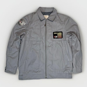 May include: A light gray zip-up jacket featuring a collar, long sleeves, and two front pockets. A circular patch is on the right shoulder, and a black rectangular patch on the left chest displays the text Corvette vette vette vette vette.
