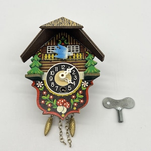May include: A wooden cuckoo clock featuring a dark brown gabled roof, a light blue bird, and two green pine trees. The clock face has white numbers on a black background with the word GERMANY printed at the bottom. Includes a metal winding key.