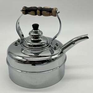 Könnte beinhalten: Ein Teekessel aus poliertem Chrom mit zylindrischem Körper und gebogenem Ausguss. Der Kessel verfügt über einen zentralen Deckel mit einem kleinen schwarzen Knopf und einen Klappgriff mit einem dekorativen, abgenutzten Holzgriff.