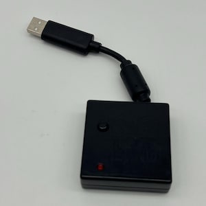 May include: A black square electronic device with a short cable ending in a USB-A connector. The device features a small circular button and a single red LED indicator light on its top surface, along with a molded logo design.