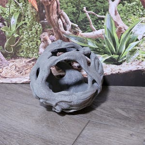 May include: A spherical, grey, textured object with an open, hollow centre and a complex, branch-like exterior structure. The surface has a rough, matte finish with multiple irregular openings throughout the form, creating a hollow, cage-like appearance.
