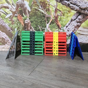 Puede incluir: Cuatro estructuras rectangulares plegables de plástico colocadas en posición vertical. Una es gris, una es verde con una franja central negra, una es roja con una franja central amarilla y una es azul. Cada una presenta un diseño de listones.
