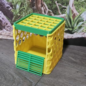 Puede incluir: Una estructura cúbica amarilla con una tapa verde de rejilla y una puerta verde de listones en la parte frontal. Las paredes laterales presentan un diseño repetitivo de recortes circulares. La estructura está hecha de plástico rígido.