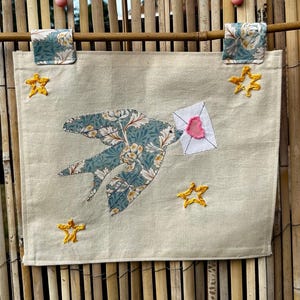 Puede incluir: Un tapiz rectangular de tela color crema con una aplicación central de un pájaro con un patrón floral azul y blanco. El pájaro lleva un sobre blanco con un corazón rosa. Cuatro estrellas amarillas bordadas rodean al pájaro.