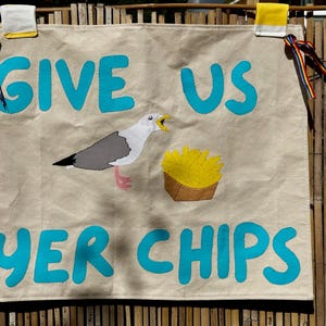 May include: A rectangular beige fabric wall hanging featuring the bright blue text GIVE US YER CHIPS. In the center, a fabric applique depicts a seagull with white, gray, and black feathers standing next to a brown paper container of yellow french fries.