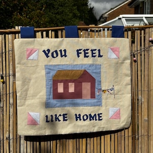 May include: A rectangular cream-coloured fabric wall hanging with three dark blue tabs at the top. It features the dark blue text "YOU FEEL LIKE HOME" surrounding a central applique of a house with a brown roof and a blue gingham background.