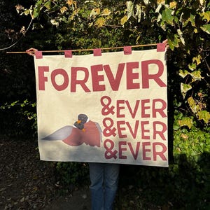 Könnte beinhalten: Ein rechteckiger cremefarbener Wandbehang aus Stoff, der an einer Holzstange mit roten Stofflaschen befestigt ist. Das Banner zeigt den dunkelroten Text FOREVER &EVER &EVER &EVER und eine grafische Illustration zweier Enten mit grauem und braunem Gefieder.