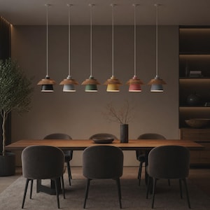 May include: Six pendant lights hang in a row above a long wooden dining table. Each light features a wooden top section and a colored base in black, light gray, green, yellow, pink, and dark gray. Six upholstered chairs are arranged around the table.